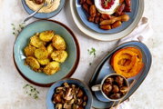 Draufsicht auf gedeckten Tisch mit blauen Tellern: gewürzte Kartoffeln, Pilze, Datteln mit Dip und Marmelade, Kürbisscheiben sowie geröstete Kastanien in einer Tasse.