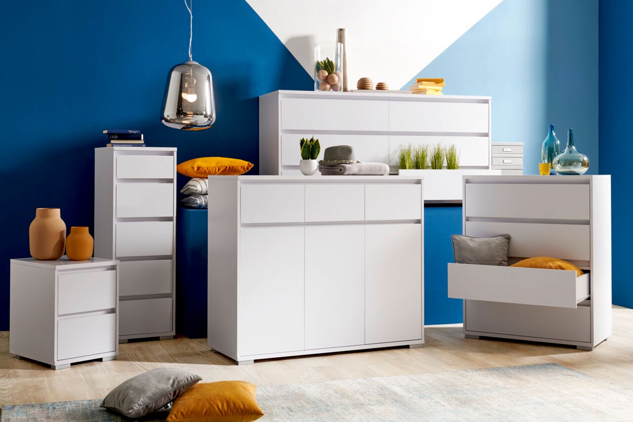 Carryhome Nachtkommode LINE Moderne weiße Möbelkollektion mit Hochkommode, Sideboard und Nachtkommode in einem stilvollen Raum mit blauer Wand, aus der Frontalperspektive.