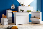 Moderne weiße Möbelkollektion mit Hochkommode, Sideboard und Nachtkommode in einem stilvollen Raum mit blauer Wand, aus der Frontalperspektive.