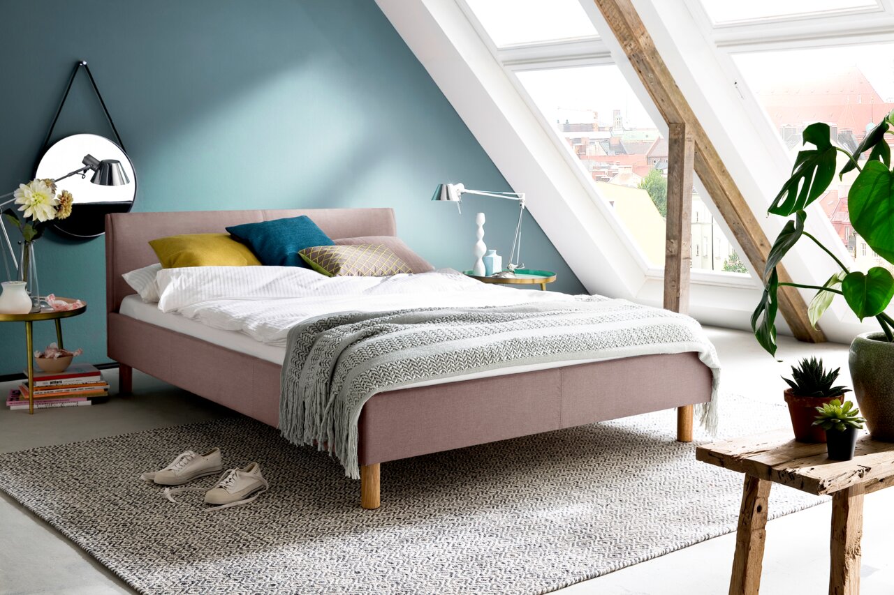 Z2 Bed met matras (120) LENA Modern gestoffeerd bed in een lichte zolderkamer met grote ramen, versierd met kleurrijke kussens en een grijze deken, zijaanzicht.