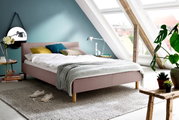 Modern gestoffeerd bed in een lichte zolderkamer met grote ramen, versierd met kleurrijke kussens en een grijze deken, zijaanzicht.
