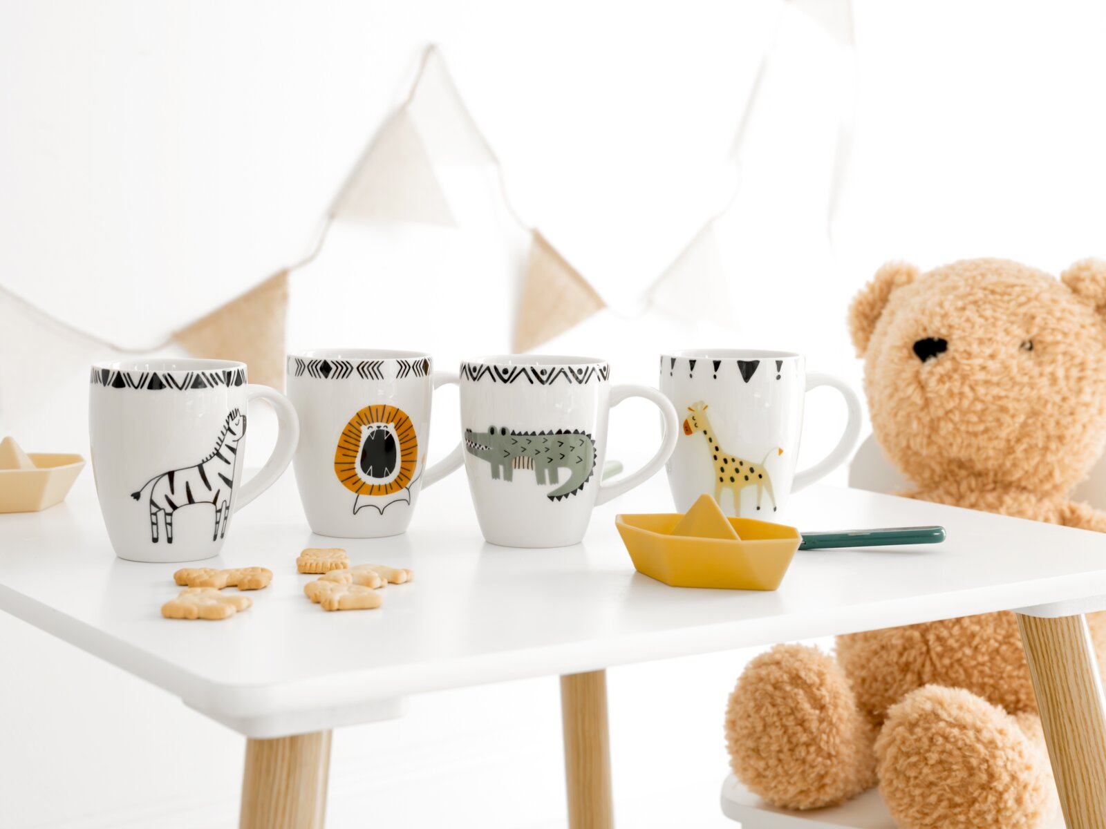 Vier weiße Kinderbecher mit Zebra-, Löwen-, Krokodil- und Giraffenmotiv in frontaler Perspektive auf einem Kindertisch, dekoriert mit Keksen, gelbem Papierboot und großem Teddybären.