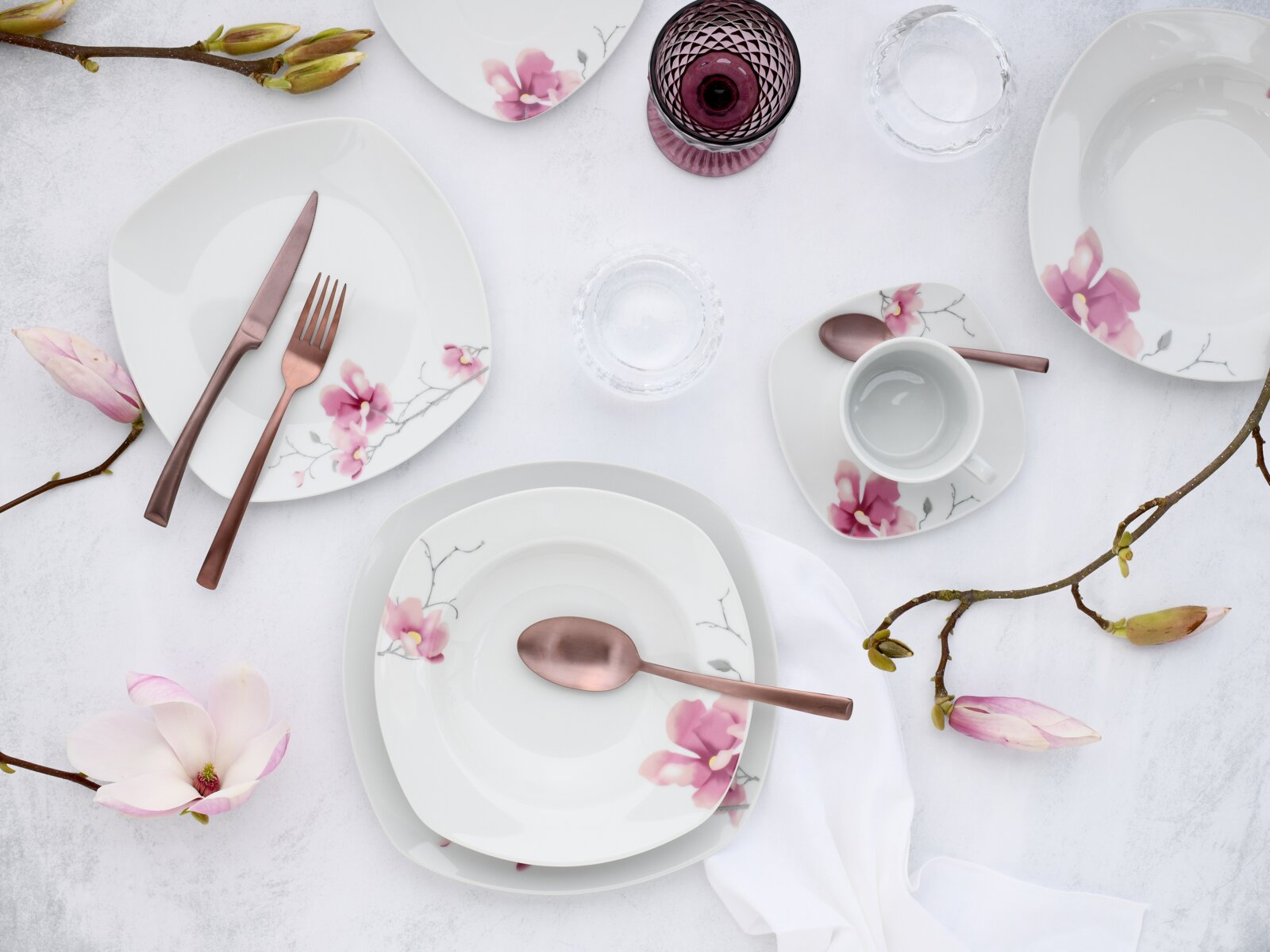 Draufsicht auf ein weißes Porzellan-Geschirrset mit rosa Blütenmotiv (Teller, Schalen, Tasse mit Untertasse) und roségoldenem Besteck auf hellem Tischarrangement mit Magnolienzweigen und Gläsern.