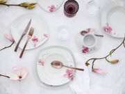 Draufsicht auf ein weißes Porzellan-Geschirrset mit rosa Blütenmotiv (Teller, Schalen, Tasse mit Untertasse) und roségoldenem Besteck auf hellem Tischarrangement mit Magnolienzweigen und Gläsern.