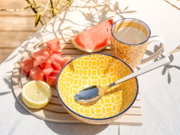 Gelbe Keramikschale mit geometrischem Muster und blauem Rand, daneben passende Tasse mit hellem Getränk, Löffel, Wassermelonenstücke und halbe Zitrone auf Holztablett im Sonnenlicht; Perspektive: leicht von oben schräg