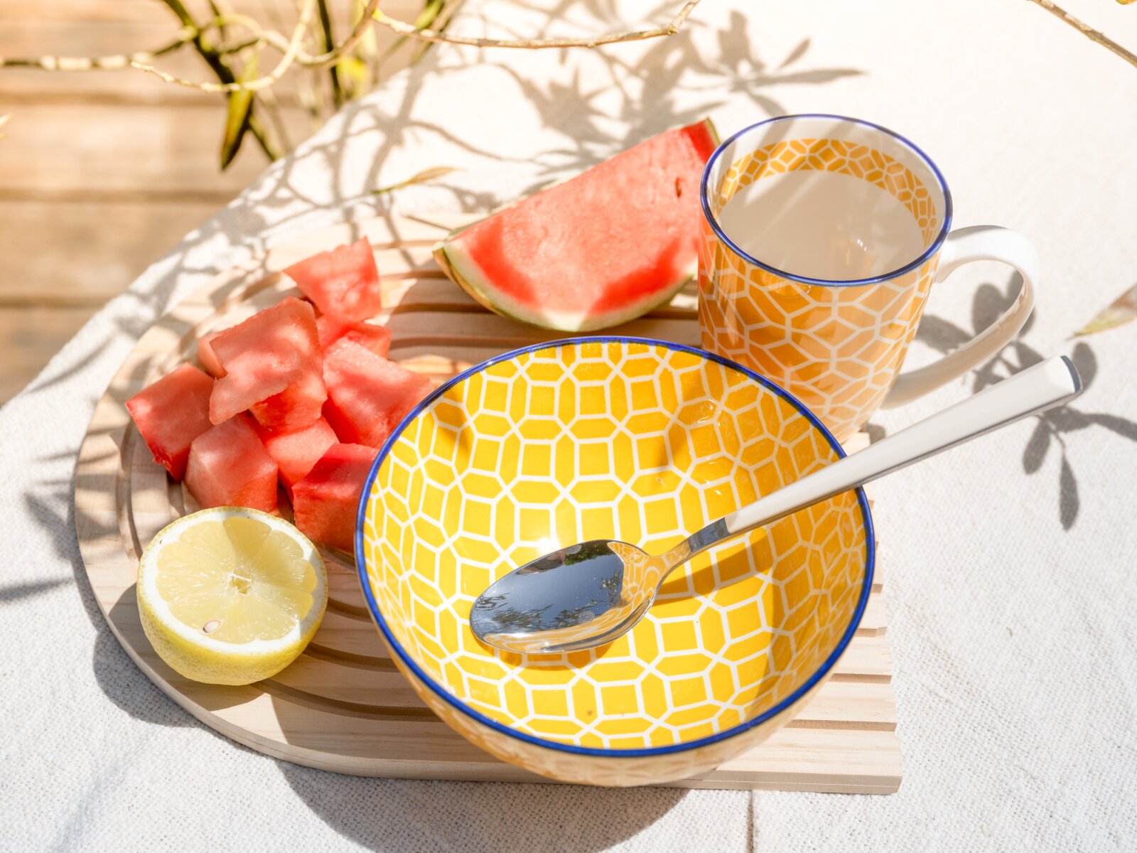 Gelbe Keramikschale mit geometrischem Muster und blauem Rand, daneben passende Tasse mit hellem Getränk, Löffel, Wassermelonenstücke und halbe Zitrone auf Holztablett im Sonnenlicht; Perspektive: leicht von oben schräg