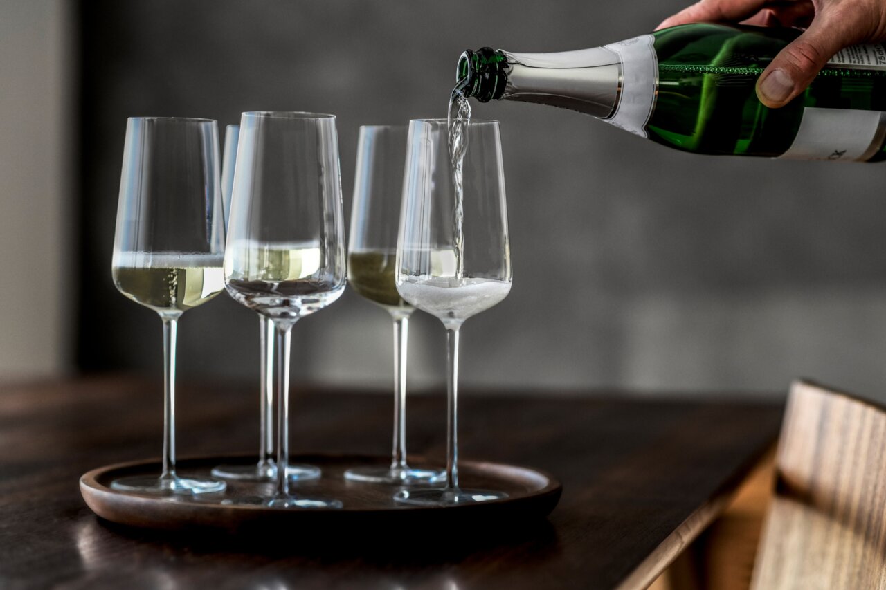 Vier champagneglazen uit de Vervino-serie op een dienblad, gezien vanuit de zijkant, terwijl er in een van de glazen champagne wordt ingeschonken.