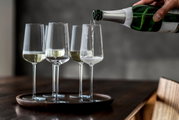 Vier Champagnergläser der Serie Vervino auf einem Tablett, aus der seitlichen Perspektive, während in eines der Gläser Champagner eingeschenkt wird.