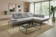 Modernes graues L-förmiges Ecksofa mit gesteppter Sitzfläche, schwarzem Metallgestell und Ottomane rechts in hellem Wohnzimmer; Perspektive: frontale leicht schräge Ansicht von links