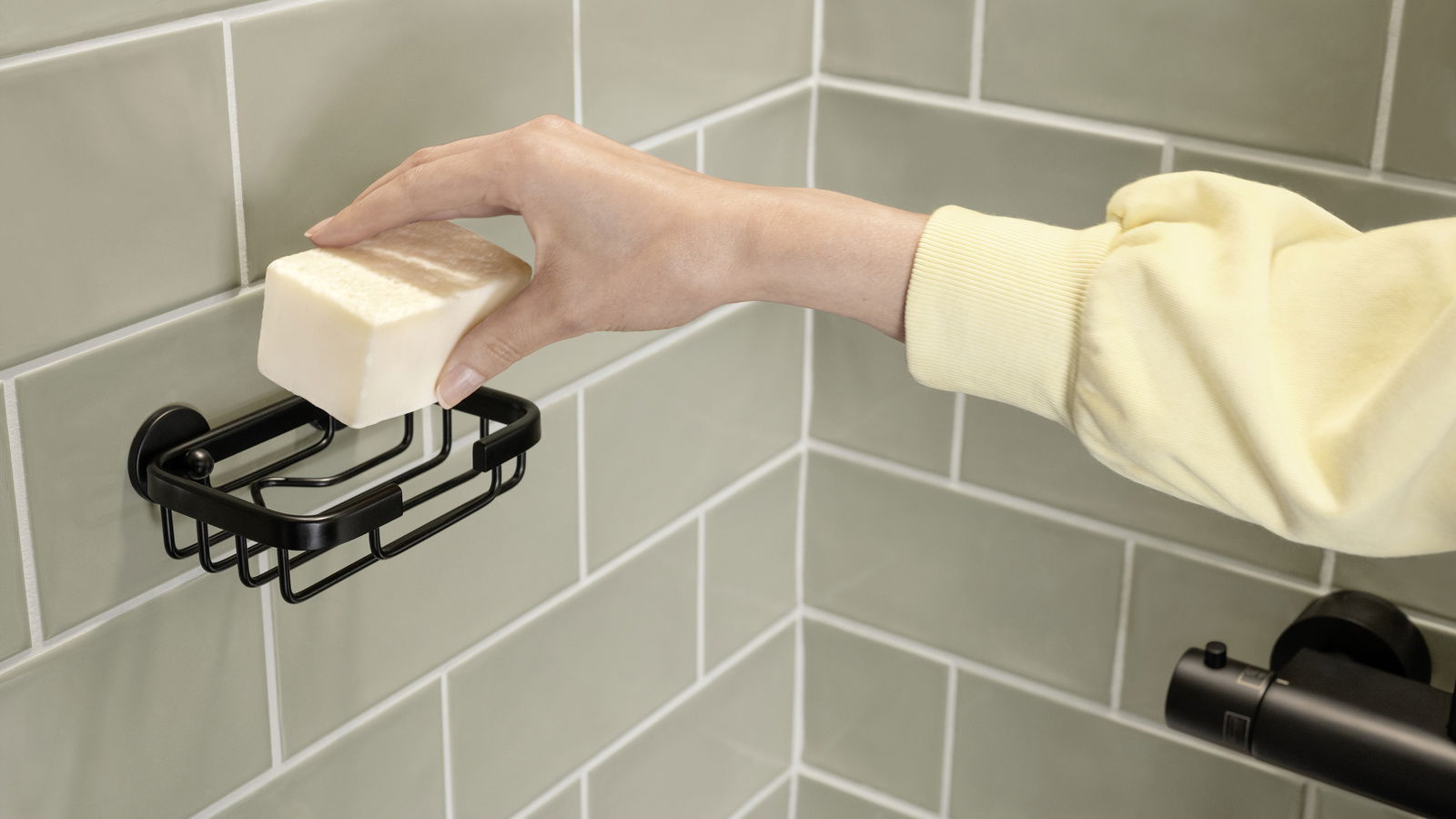 Hand legt ein Stück Seife in eine schwarze Seifenschale an der Wand, seitliche Perspektive