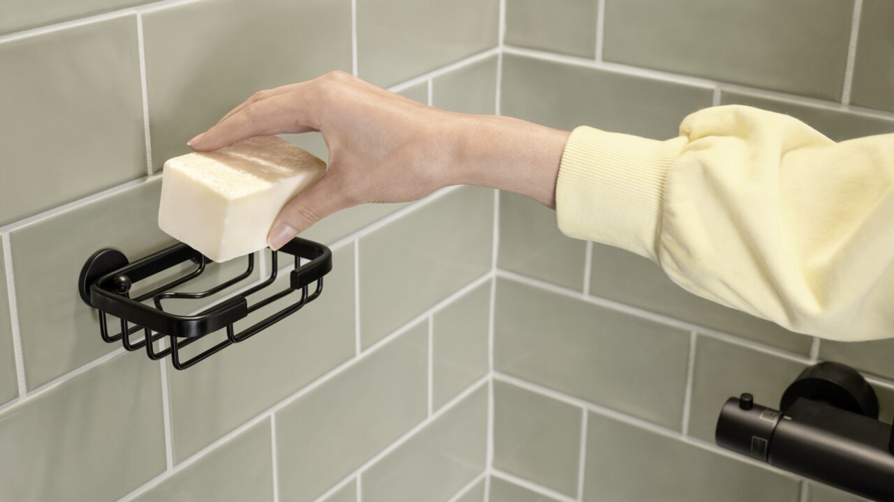 Tesa Seifenschale ALUXX BLACK Hand legt ein Stück Seife in eine schwarze Seifenschale an der Wand, seitliche Perspektive