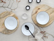 Draufsicht auf einen gedeckten Tisch mit weißen Schalen mit schwarzem Rand auf runden Holzplatten, schwarzem Löffel, kleinen schwarzen Teelichtern, Glas und dekorativen Zweigen auf hellem Leinentuch.