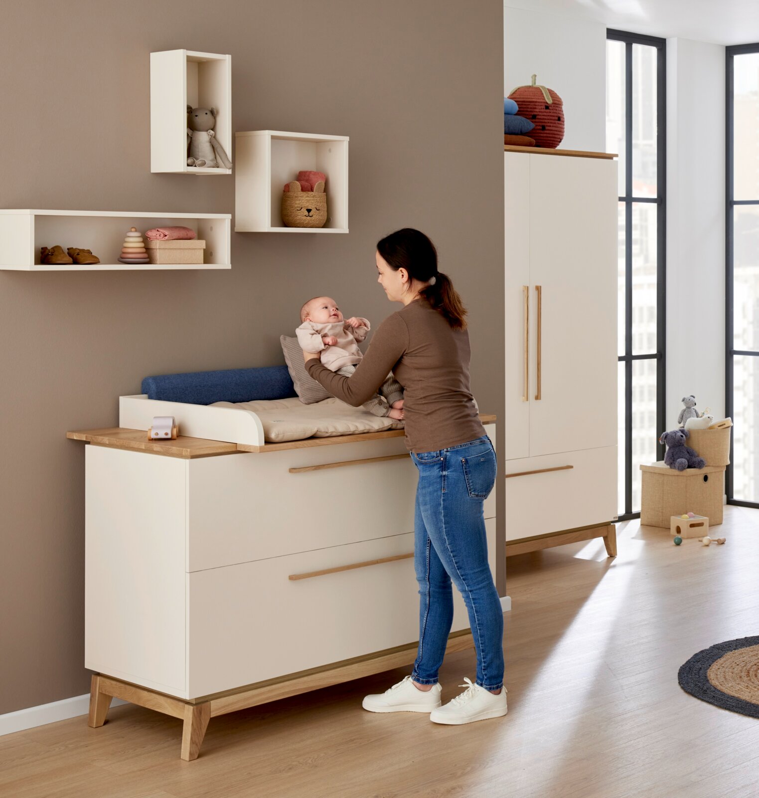 Frau steht vor einer Wickelkommode und hält ein Baby, im Hintergrund sind Wandboxen an der Wand montiert. Perspektive von der Seite.