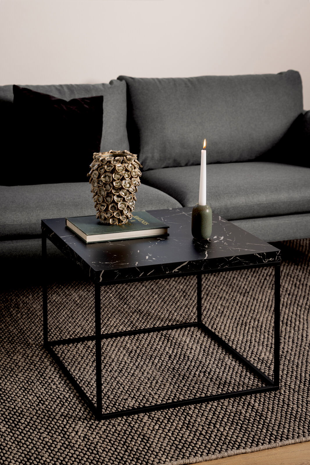 Moderner Couchtisch mit schwarzer Marmoroptik, dekoriert mit einer Kerze und einem Buch, vor einem grauen Sofa, aus seitlicher Perspektive.
