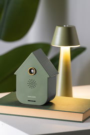 Grüne Soundbox in Form eines Hauses, frontal fotografiert, auf einem Buch stehend, mit einer Lampe im Hintergrund.