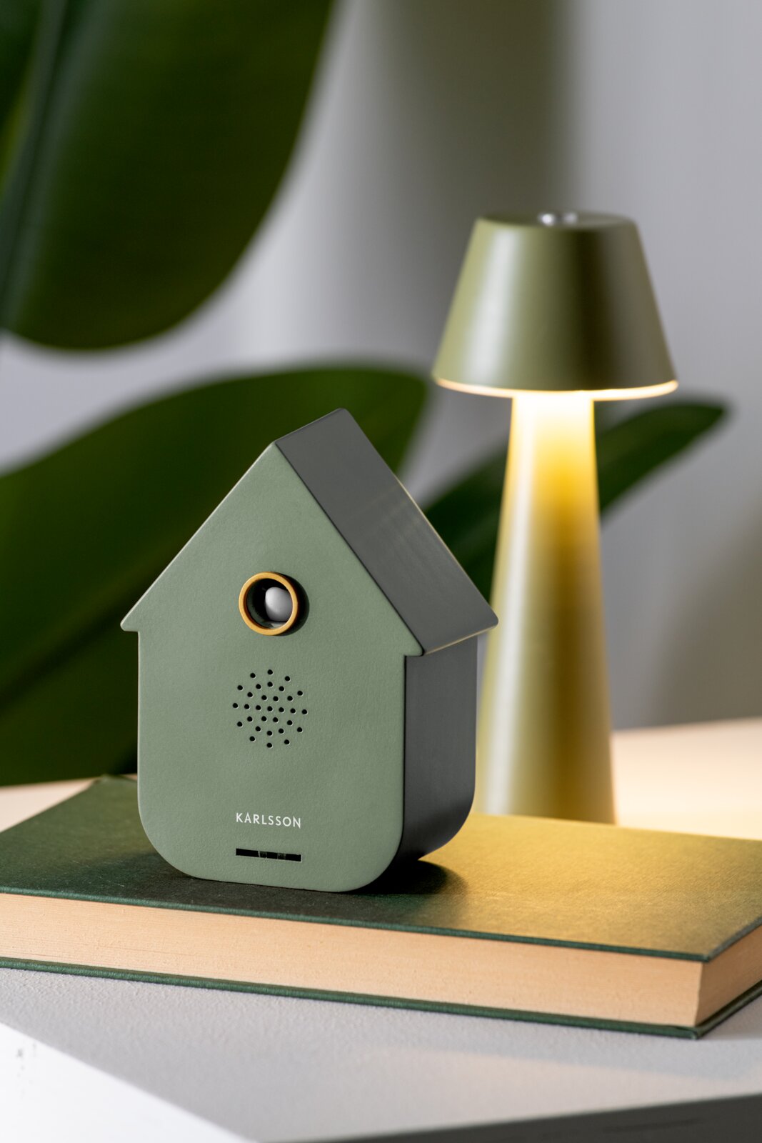 KARLSSON Soundbox CUCKOO Grüne Soundbox in Form eines Hauses, frontal fotografiert, auf einem Buch stehend, mit einer Lampe im Hintergrund.