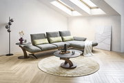 Modernes graues Ecksofa mit verstellbaren Rückenlehnen in hellem Wohnzimmer, davor runder Couchtisch auf Teppich; Perspektive: frontale leicht schräge Ansicht von links.