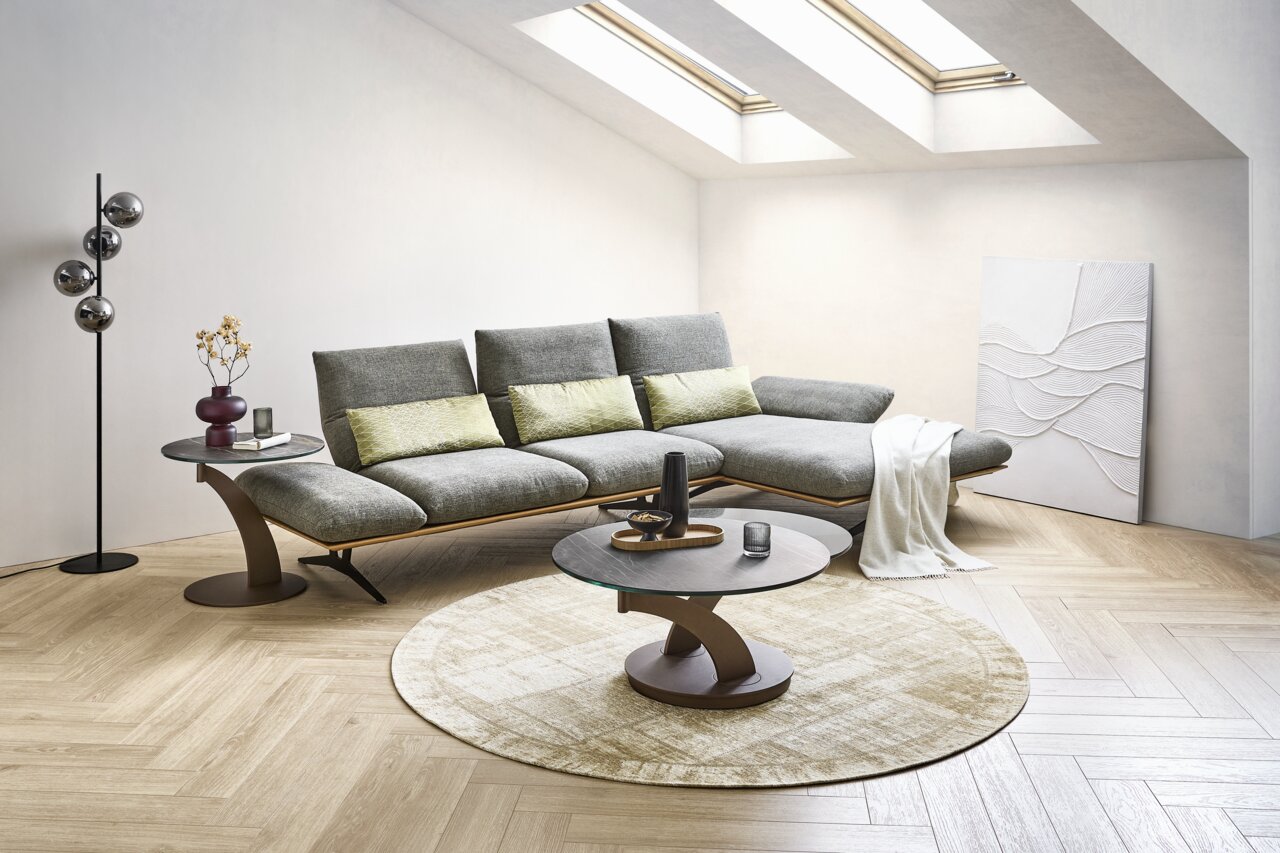 Modernes graues Ecksofa mit verstellbaren Rückenlehnen in hellem Wohnzimmer, davor runder Couchtisch auf Teppich; Perspektive: frontale leicht schräge Ansicht von links.