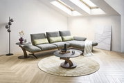 Modernes graues Ecksofa mit verstellbaren Rückenlehnen in hellem Wohnzimmer, davor runder Couchtisch auf Teppich; Perspektive: frontale leicht schräge Ansicht von links.