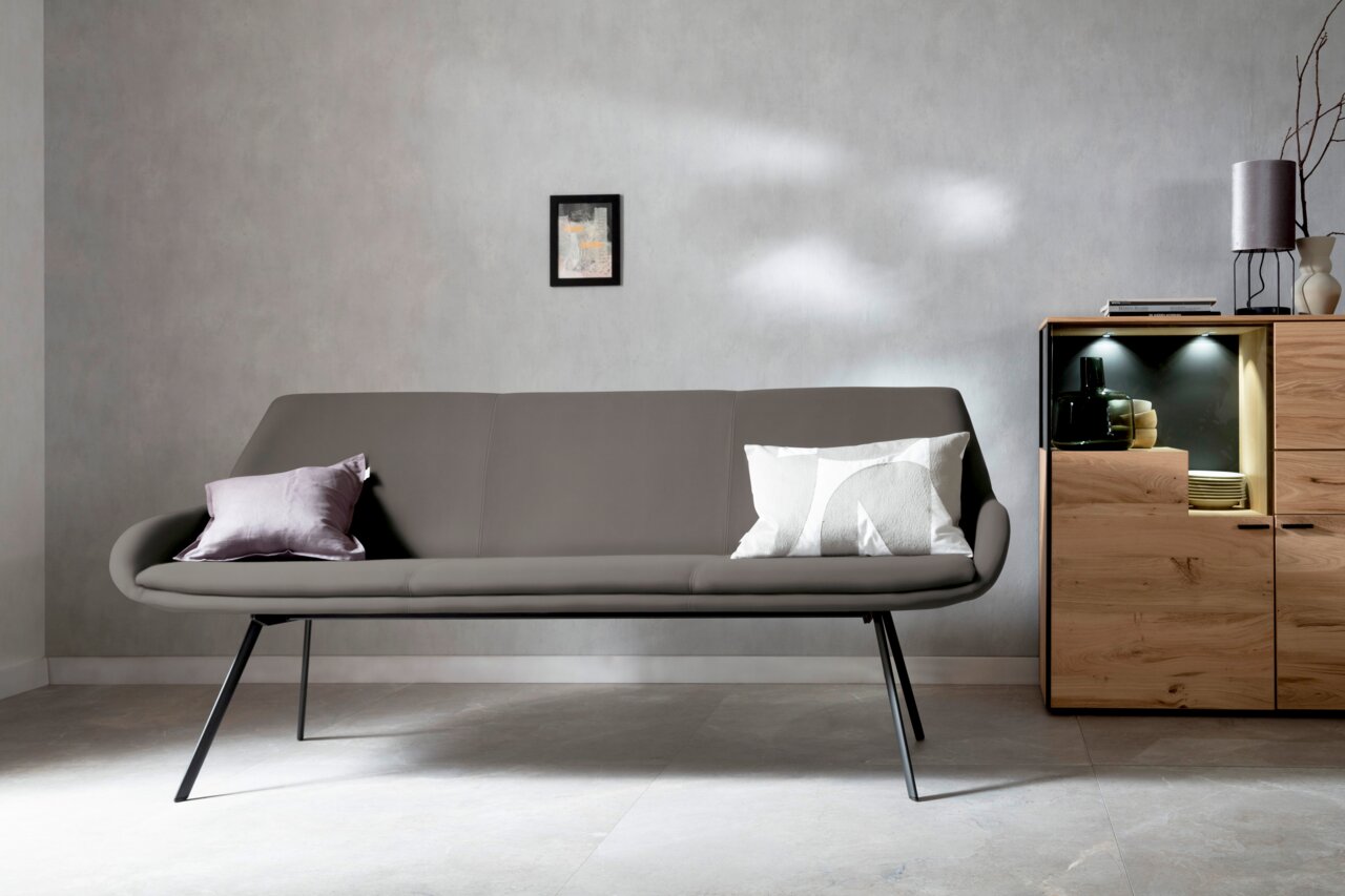Solitärbank 3er in modernem Design, frontal fotografiert, mit grauem Bezug und zwei dekorativen Kissen, in einem stilvollen Raum mit Holzmöbeln.