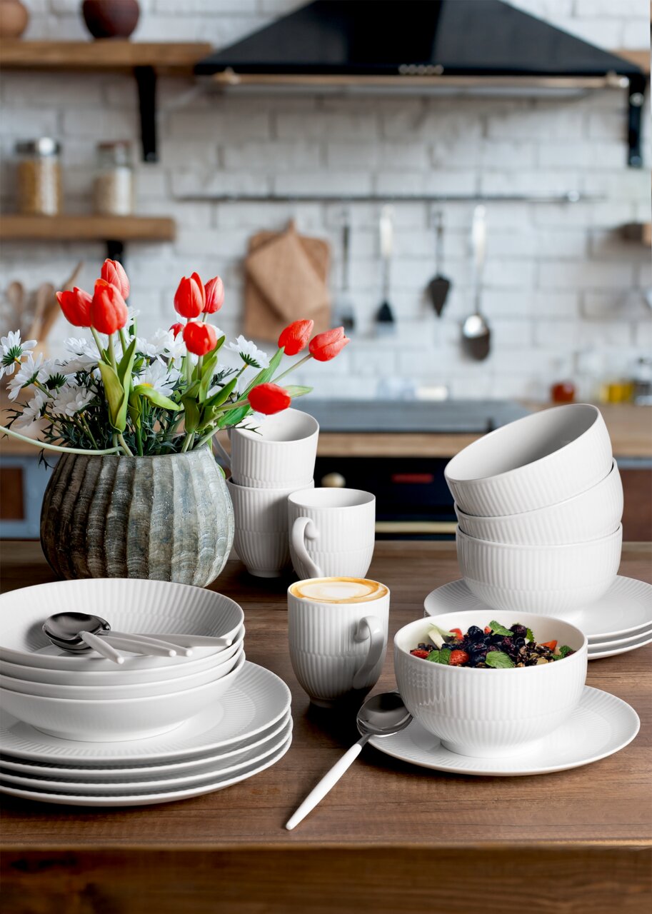 30-delige servieset in een moderne keuken, bestaande uit witte borden, schalen en kopjes, met een bloemstuk op de achtergrond. Perspectief: vooraanzicht van de servieset.