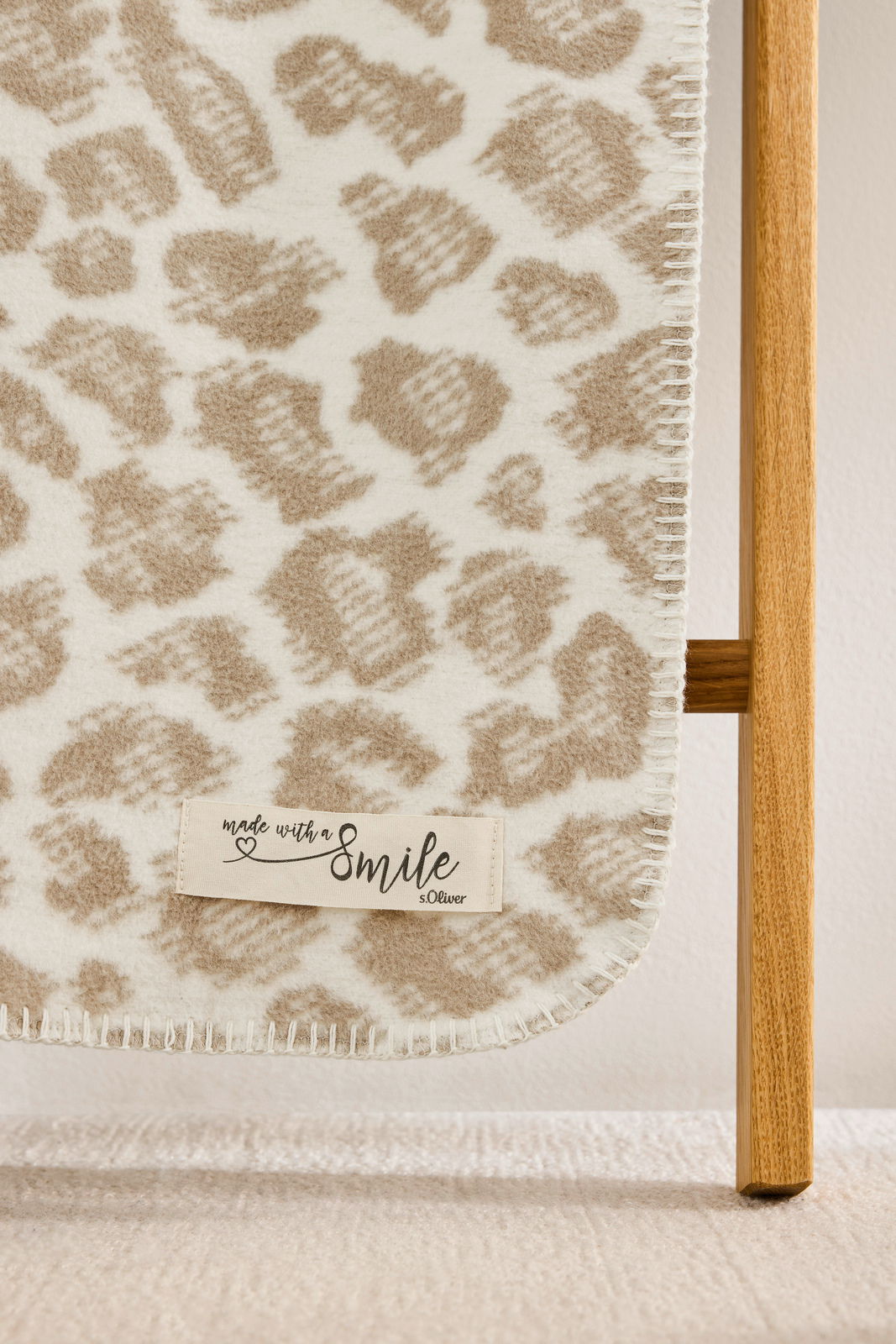 Nahaufnahme einer beige-weißen Wohndecke mit Leopardenmuster, aufgehängt an einem Holzgestell, mit einem Etikett 'made with a smile s.Oliver'.