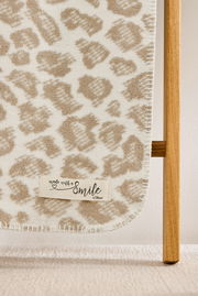 Nahaufnahme einer beige-weißen Wohndecke mit Leopardenmuster, aufgehängt an einem Holzgestell, mit einem Etikett 'made with a smile s.Oliver'.