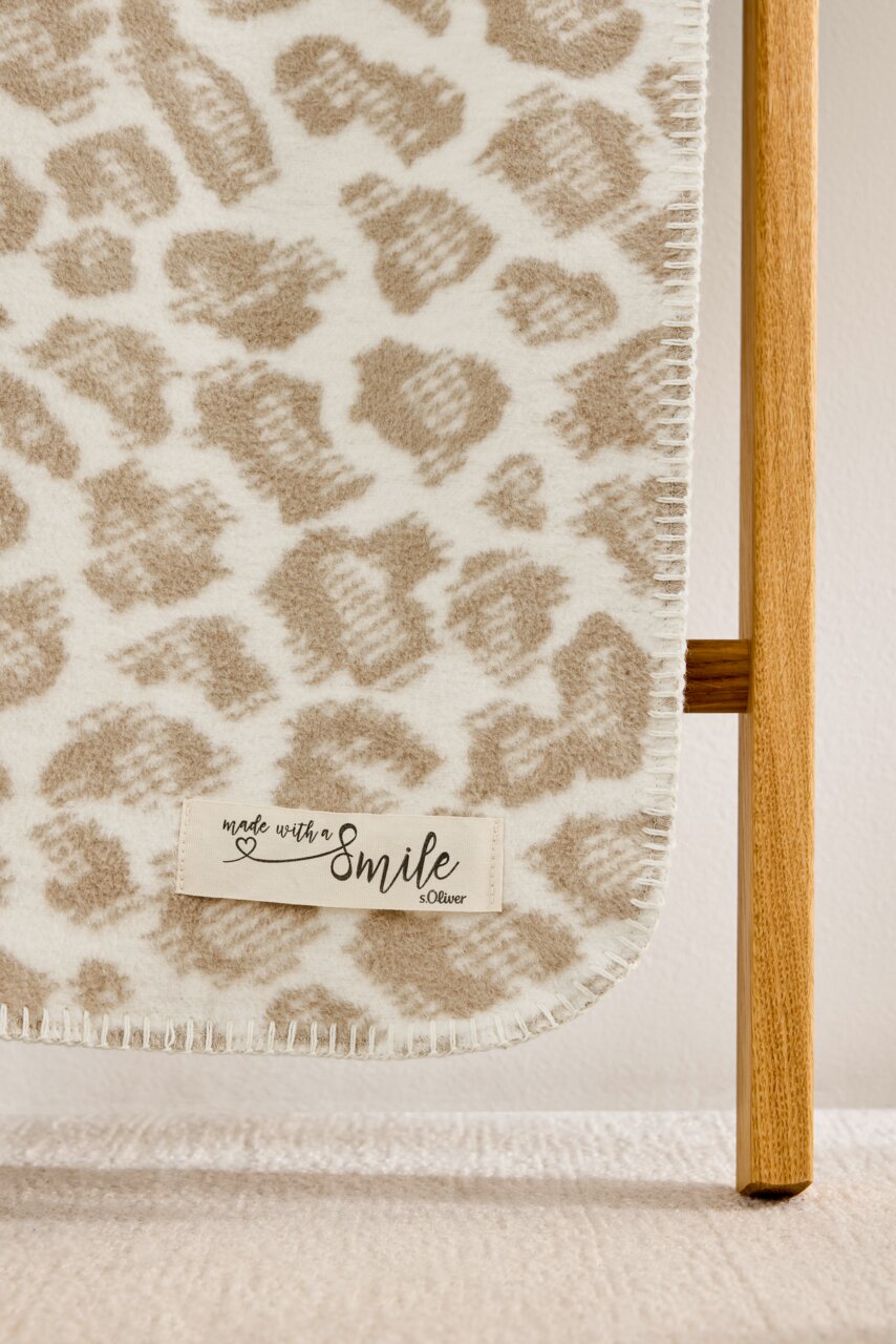 Nahaufnahme einer beige-weißen Wohndecke mit Leopardenmuster, aufgehängt an einem Holzgestell, mit einem Etikett 'made with a smile s.Oliver'.