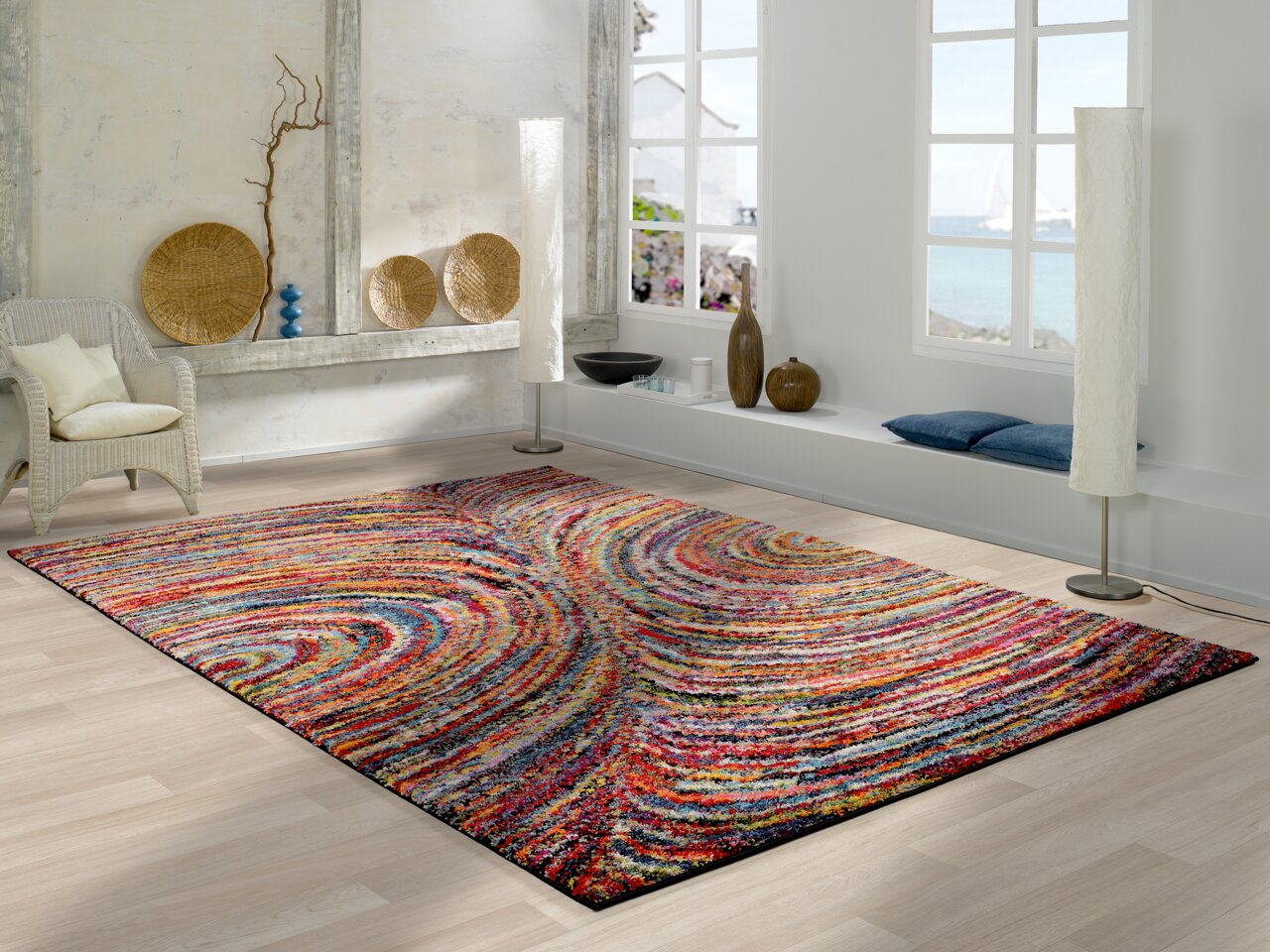 OCI Webteppich 120x170 cm SIXTEEN Bunter Teppich mit wellenförmigem Muster in einem modernen Wohnzimmer mit Blick auf das Meer durch große Fenster.