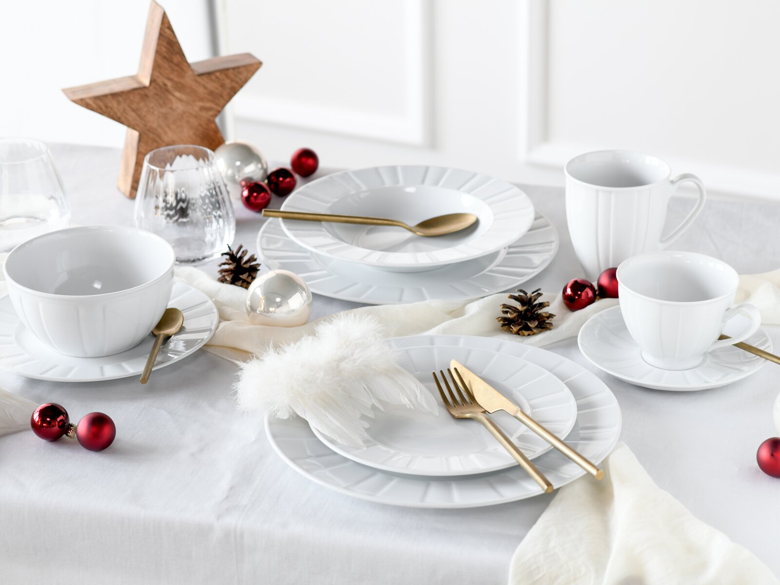 Festlich gedeckter Tisch mit weißem Porzellan-Geschirrset (Teller, Schalen, Tassen), goldfarbenem Besteck und Weihnachtsdeko in Rot/Silber, aufgenommen in leicht schräger Seitenansicht von oben.
