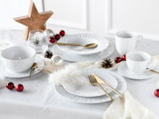 Festlich gedeckter Tisch mit weißem Porzellan-Geschirrset (Teller, Schalen, Tassen), goldfarbenem Besteck und Weihnachtsdeko in Rot/Silber, aufgenommen in leicht schräger Seitenansicht von oben.