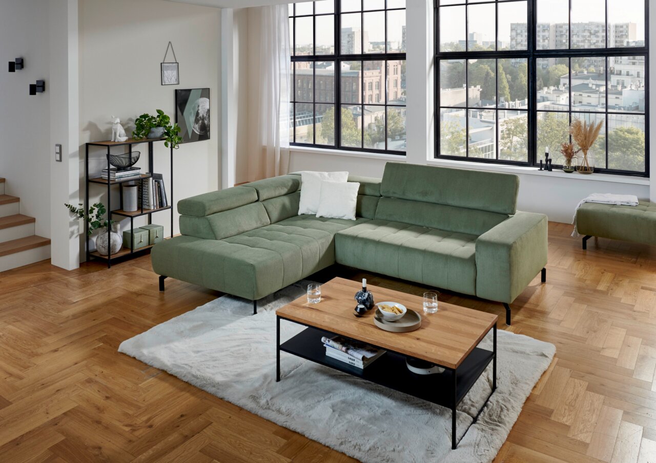 Grüne Eckgarnitur in modernem Wohnzimmer mit großen Fenstern, Holzboden und einem Couchtisch, Perspektive von vorne links.