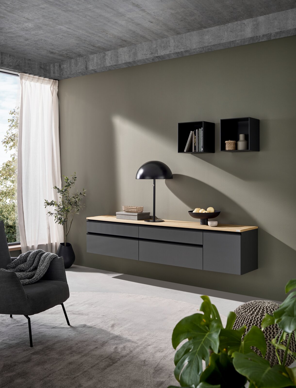 Modernes, schwebendes Sideboard in Anthrazit mit heller Holzplatte an olivgrüner Wand, dekoriert mit Tischlampe und Schale; perspektivische Ansicht von schräg vorn/leicht seitlich im Wohnzimmer.