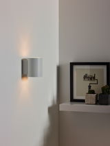 Moderne Wandleuchte in zylindrischer Form, seitliche Perspektive, montiert an einer weißen Wand neben einem Regal mit Dekoration.