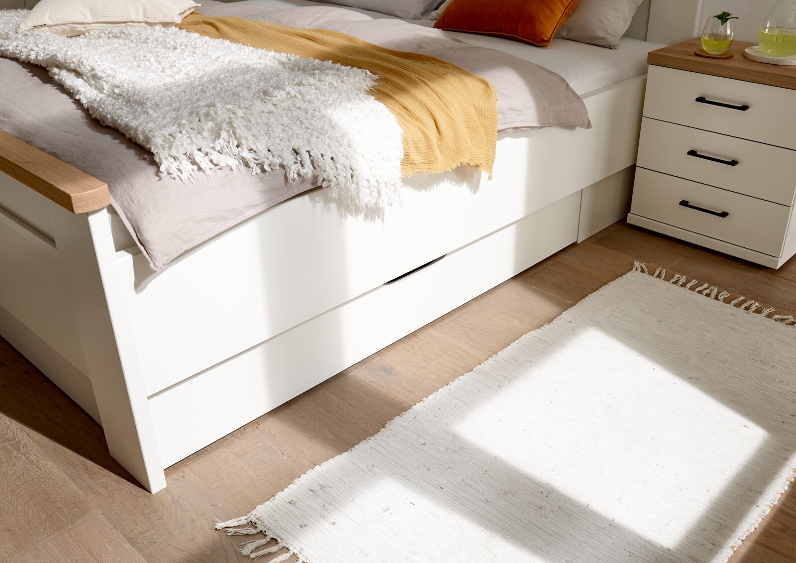 Modernes weißes Bett mit Schubladen und Holzarmlehne, dekoriert mit gelber Decke und weißem Plaid; daneben Nachttisch und heller Teppich – Perspektive: seitliche Nahaufnahme auf Bettseite und Stauraum.