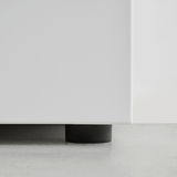 Nahaufnahme der unteren Ecke einer weißen Kommode mit schwarzem Fuß auf grauem Boden.