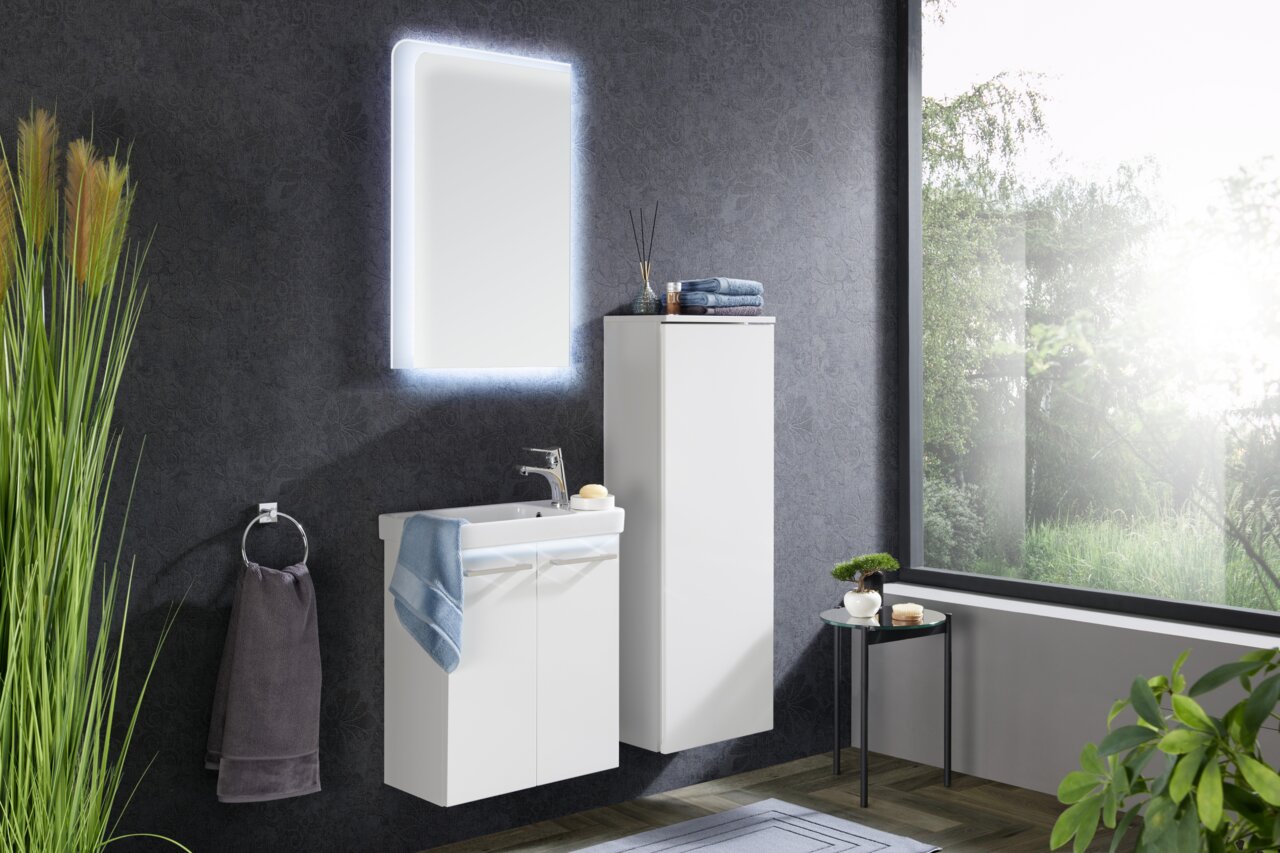 Modernes Badezimmer mit beleuchtetem Spiegelelement, Waschbeckenunterschrank und hohem Schrank in weißer Hochglanzoptik, seitliche Perspektive.