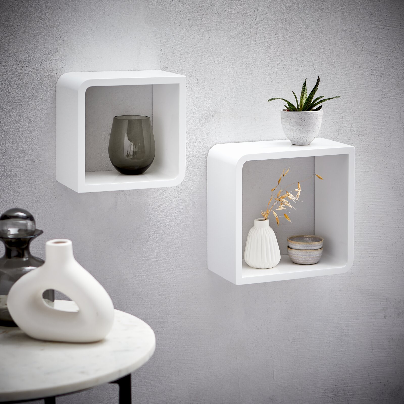 XORA Set van 2 wandplanken DUO Zwei weiße, quadratische Wandregale aus Holz, an einer grauen Wand montiert, mit dekorativen Vasen und Pflanzen, seitliche Perspektive.