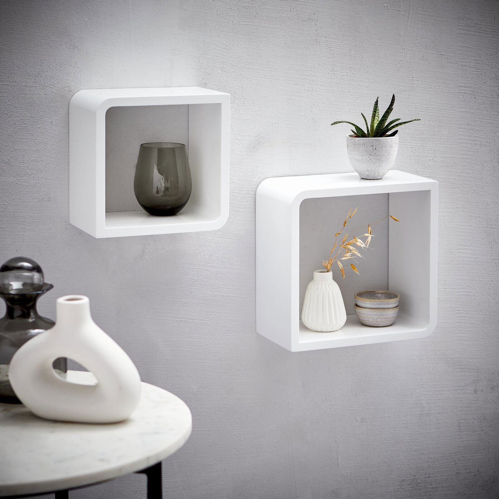 Zwei weiße, quadratische Wandregale aus Holz, an einer grauen Wand montiert, mit dekorativen Vasen und Pflanzen, seitliche Perspektive.