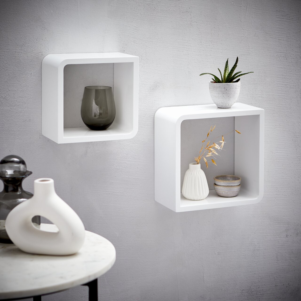 Zwei weiße, quadratische Wandregale aus Holz, an einer grauen Wand montiert, mit dekorativen Vasen und Pflanzen, seitliche Perspektive.
