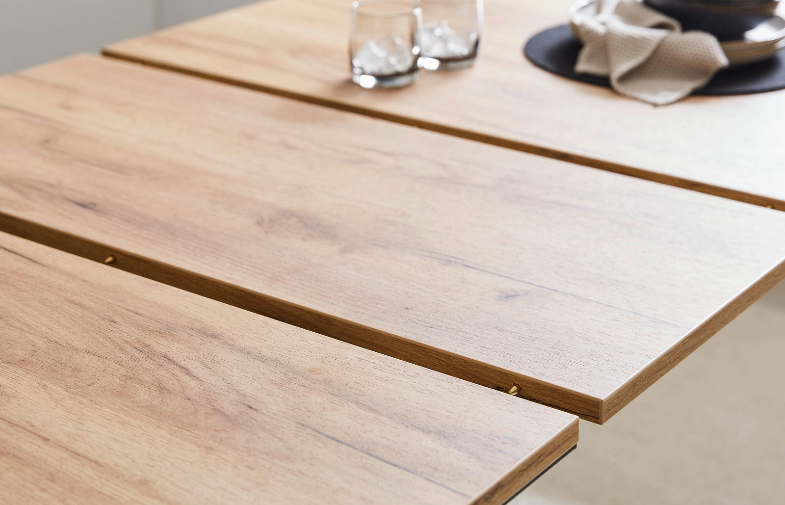 Nahaufnahme eines Esstisches aus Holz mit natürlicher Maserung, darauf zwei Gläser mit Eiswürfeln und ein gedeckter Platz mit Serviette, aus seitlicher Perspektive.