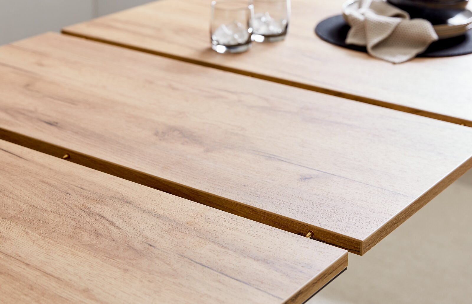Nahaufnahme eines Esstisches aus Holz mit natürlicher Maserung, darauf zwei Gläser mit Eiswürfeln und ein gedeckter Platz mit Serviette, aus seitlicher Perspektive.