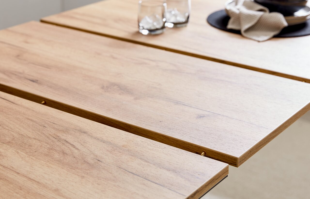 Nahaufnahme eines Esstisches aus Holz mit natürlicher Maserung, darauf zwei Gläser mit Eiswürfeln und ein gedeckter Platz mit Serviette, aus seitlicher Perspektive.
