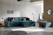 Modernes blaugrünes Ecksofa mit Récamiere und passendem Hocker in einem hellen Wohnzimmer, Frontansicht in leichter Schrägperspektive von links