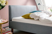 Zijaanzicht van een grijs gestoffeerd bed met hoofdeinde, versierd met kleurrijke kussens en wit beddengoed, tegen een roze muur.