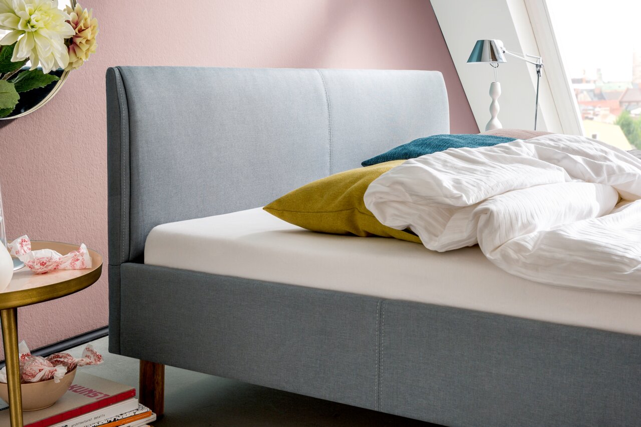 Zijaanzicht van een grijs gestoffeerd bed met hoofdeinde, versierd met kleurrijke kussens en wit beddengoed, tegen een roze muur.