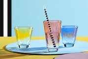 Drie kleurrijke drinkglazen, waarvan één rood met een gestreept rietje, gezien van voren tegen een kleurrijke achtergrond.