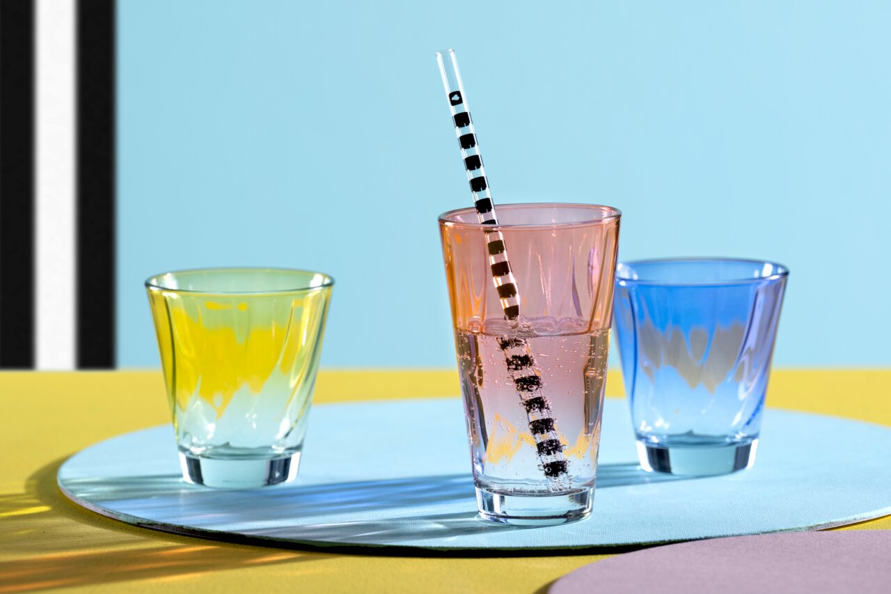 Drie kleurrijke drinkglazen, waarvan één rood met een gestreept rietje, gezien van voren tegen een kleurrijke achtergrond.