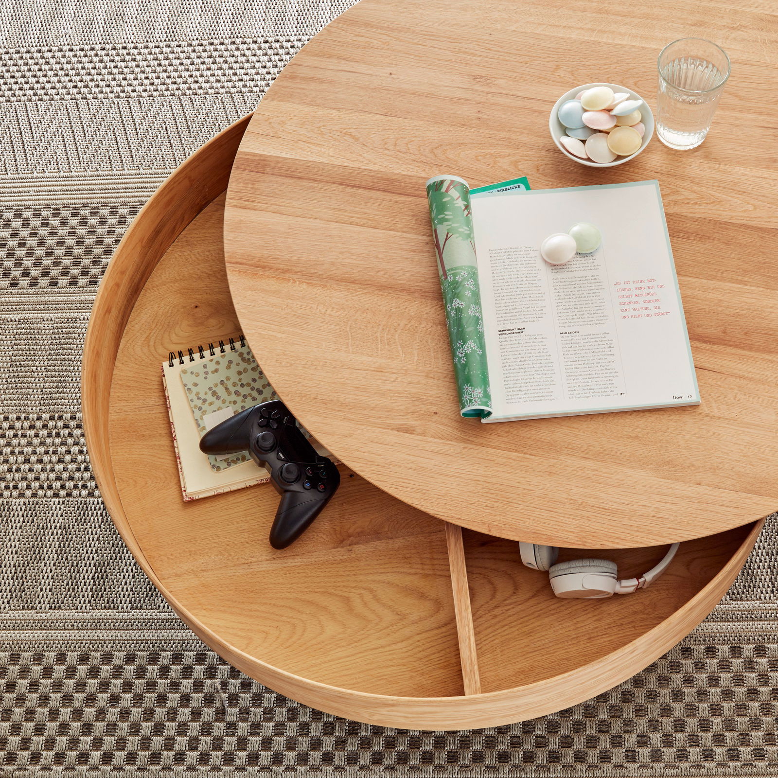 Runder Couchtisch aus Holz mit drehbarer Tischplatte, darunter Stauraum mit Kopfhörern und Notizbuch, von oben betrachtet.