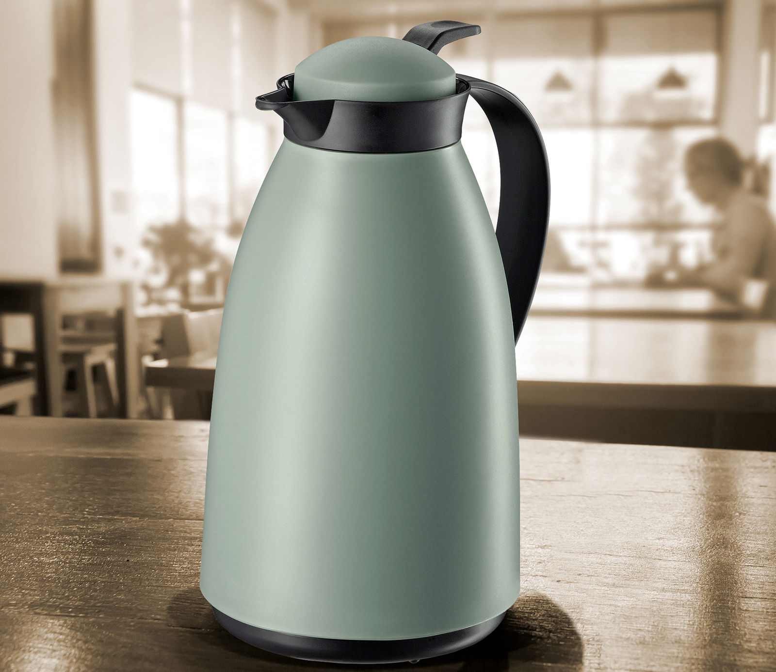 Grüne matte Isolierkanne mit schwarzem Griff und Deckel, auf einem Tisch in einem Café aus seitlicher Perspektive.